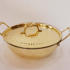 May include: A gold-coloured cooking pot with a matching lid. The pot has two handles and a lid handle. The pot and lid have a hammered texture. The pot is round and sits on a white surface.