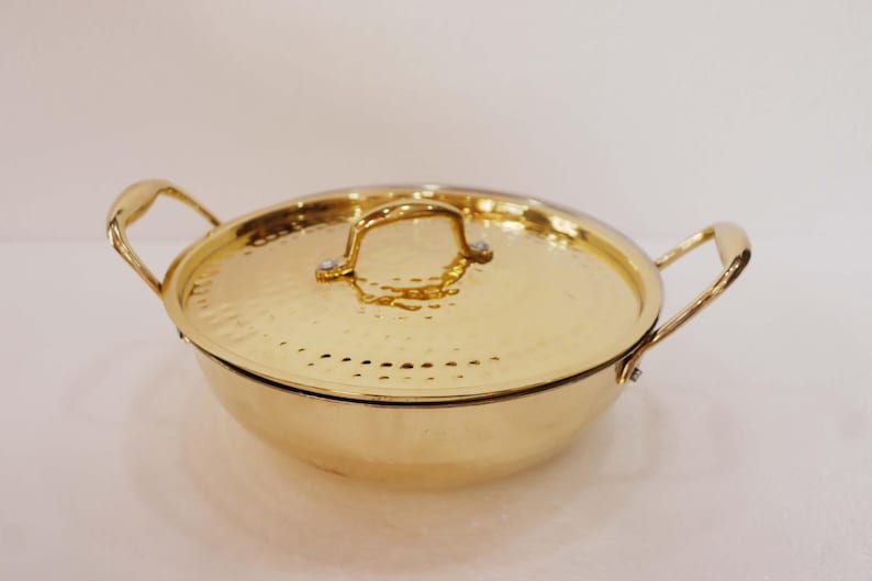 May include: A gold-coloured cooking pot with a matching lid. The pot has two handles and a lid handle. The pot and lid have a hammered texture. The pot is round and sits on a white surface.