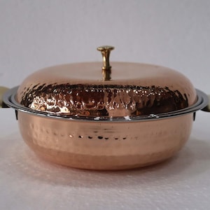 Puede incluir: Un plato de servir de color cobre con una textura martillada, con tapa y dos asas laterales. La tapa está rematada con un pomo dorado. El plato tiene un borde plateado y está diseñado para servir alimentos.