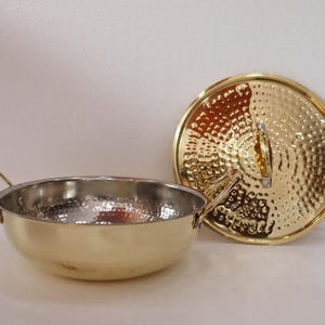 May include: A gold-coloured cooking pot made of hammered metal, with two handles and a matching lid. The pot has a shiny, silver interior. The lid is round with a textured surface and a small handle.