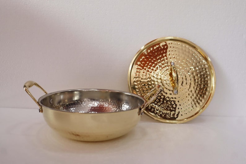 May include: A gold-coloured cooking pot made of hammered metal, with two handles and a matching lid. The pot has a shiny, silver interior. The lid is round with a textured surface and a small handle.