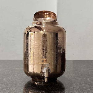 May include: A copper-coloured, ornate water dispenser with a tap and a lid. The dispenser has an engraved floral pattern and a rounded shape. The lid is open, revealing the interior. The dispenser sits on a dark surface.