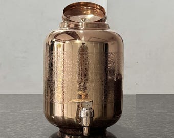 Könnte beinhalten: Ein kupferfarbener, verzierter Wasserspender mit Zapfhahn und Deckel. Der Spender hat ein eingraviertes Blumenmuster und eine abgerundete Form. Der Deckel ist geöffnet und gibt den Blick ins Innere frei. Der Spender steht auf einer dunklen Oberfläche.