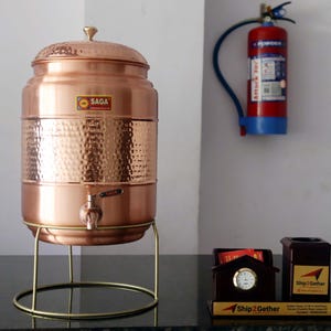 May include: A copper water dispenser with a hammered texture, a spigot, and a lid. It sits on a gold-colored metal stand. The dispenser has a "SAGA" sticker. A red fire extinguisher hangs on the wall behind it.