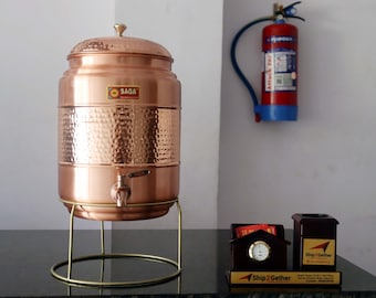 Può includere: Un distributore d'acqua in rame con una texture martellata, un rubinetto e un coperchio. È posizionato su un supporto in metallo dorato. Il distributore ha un adesivo "SAGA". Un estintore rosso è appeso al muro dietro.