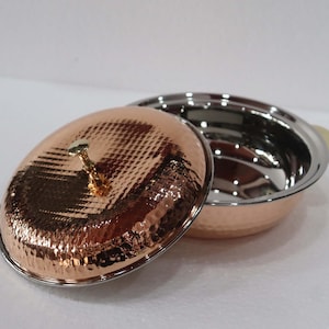 May include: A copper-colored serving dish with a hammered texture and a stainless steel interior. The lid is off to the side, revealing the inside of the dish. The dish has a gold-colored handle and a gold-colored knob on the lid.