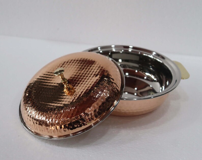 May include: A copper-colored serving dish with a hammered texture and a stainless steel interior. The lid is off to the side, revealing the inside of the dish. The dish has a gold-colored handle and a gold-colored knob on the lid.