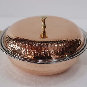 May include: A copper serving dish with a hammered texture, a lid, and brass handles. The lid is topped with a decorative brass handle. The dish has a silver rim and is designed for serving food.