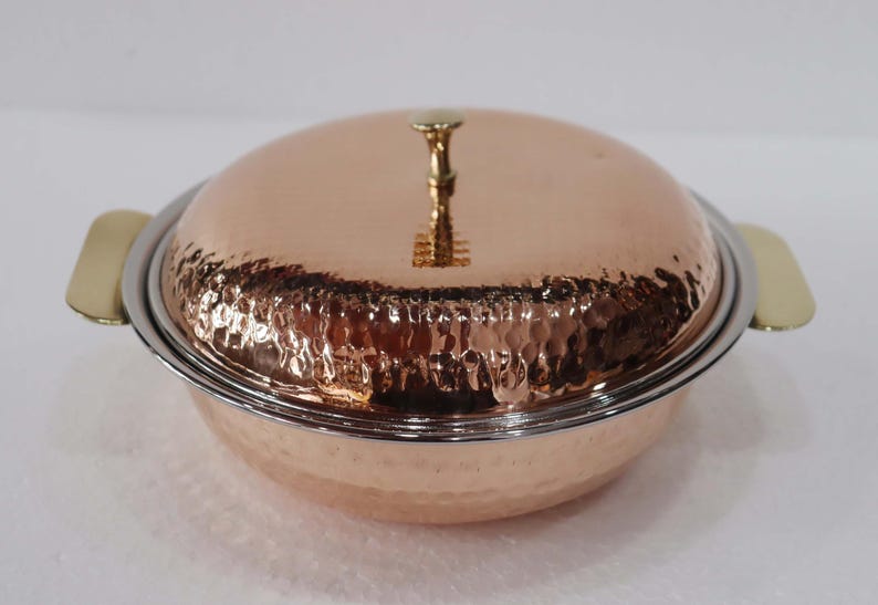 May include: A copper serving dish with a hammered texture, a lid, and brass handles. The lid is topped with a decorative brass handle. The dish has a silver rim and is designed for serving food.