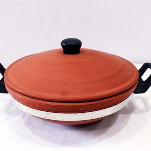 Puede incluir: Una olla de cocina de terracota con tapa y dos asas negras. La olla tiene una banda blanca con motas grises alrededor de la base. La tapa tiene un pomo negro. Esta olla de barro es adecuada para cocinar.