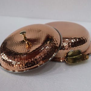 May include: Two copper-colored serving bowls with lids. The bowls have a hammered texture and a shiny finish. One lid is open, revealing the interior. The lids have a small, decorative handle. The bowls are on a white surface.