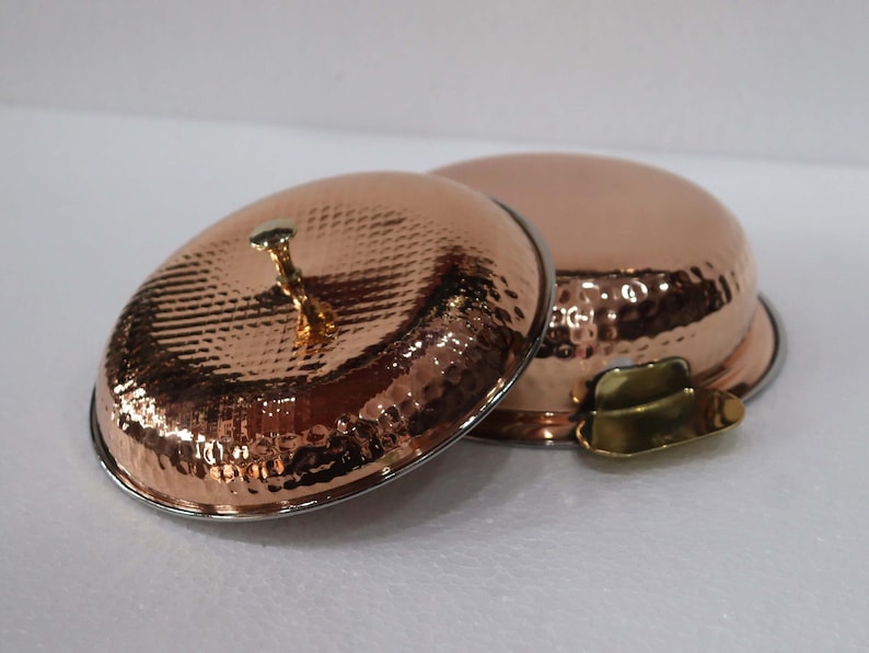 May include: Two copper-colored serving bowls with lids. The bowls have a hammered texture and a shiny finish. One lid is open, revealing the interior. The lids have a small, decorative handle. The bowls are on a white surface.