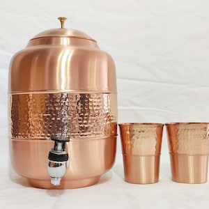 Puede incluir: Un dispensador de agua de cobre martillado con un grifo y dos vasos de cobre a juego. El dispensador tiene un diseño tradicional y es perfecto para servir agua u otras bebidas.