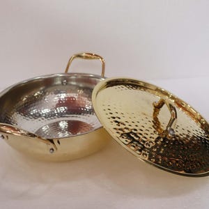 May include: A hammered metal wok with a gold exterior and a silver interior. The wok has two small handles and a larger handle on the lid. The lid is off to the side, revealing the interior of the wok.