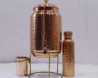 May include: A copper water dispenser with a hammered texture, a matching bottle, and a small cup. The dispenser has a spigot and sits on a brass stand. The bottle is approximately 10 inches tall.