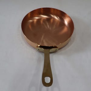 Op de afbeelding: Een koperen koekenpan met een messing handvat. De pan is rond en heeft een glanzend, reflecterend oppervlak.