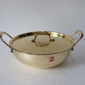 May include: A gold-coloured cooking pot with a matching lid. The pot has two handles and a lid handle. The lid and pot have a textured surface. A red and white sticker with the number 2 is on the side.
