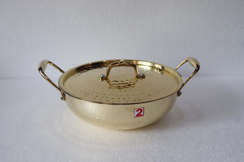 May include: A gold-coloured cooking pot with a matching lid. The pot has two handles and a lid handle. The lid and pot have a textured surface. A red and white sticker with the number 2 is on the side.