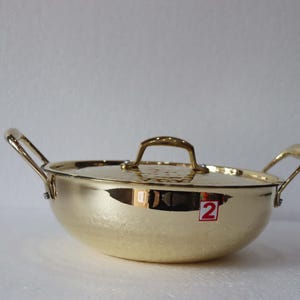 May include: A gold-coloured cooking pot with a matching lid and two side handles. The pot has a rounded shape and a textured surface. The lid has a handle on top. A red and white sticker with the number 2 is visible on the side.