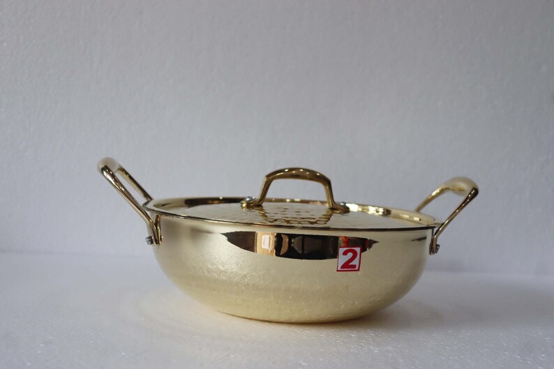 May include: A gold-coloured cooking pot with a matching lid and two side handles. The pot has a rounded shape and a textured surface. The lid has a handle on top. A red and white sticker with the number 2 is visible on the side.