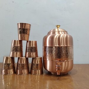 May include: A set of six copper drinking glasses and a copper water dispenser with a hammered finish. The dispenser has a spout and a gold-coloured top.