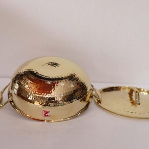 May include: A gold-coloured, hammered metal bowl with two handles and a matching lid. The bowl and lid have a textured surface and a shiny finish. The bowl is round and the lid is oval.