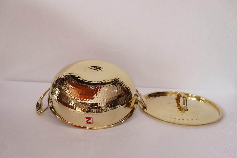 May include: A gold-coloured, hammered metal bowl with two handles and a matching lid. The bowl and lid have a textured surface and a shiny finish. The bowl is round and the lid is oval.