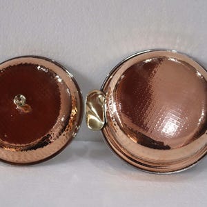 May include: Two copper-colored, round serving dishes with hammered textures. One has a lid with a small knob, the other has two gold-colored handles. The dishes are on a white surface.