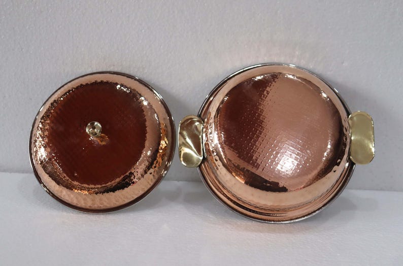May include: Two copper-colored, round serving dishes with hammered textures. One has a lid with a small knob, the other has two gold-colored handles. The dishes are on a white surface.