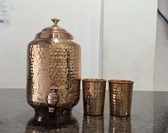 Può includere: Un distributore d'acqua in rame con una texture martellata, un rubinetto e un coperchio con un pomello dorato. Due bicchieri in rame abbinati sono accanto al distributore. Il set è su una superficie scura.