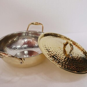 May include: A hammered metal wok with a gold-coloured rim and handles, and a matching lid. The wok's interior is silver, and the lid is off to the side. The handles are curved and the lid has a textured surface.