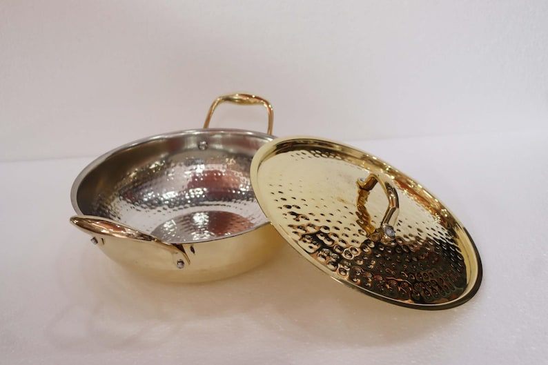 May include: A hammered metal wok with a gold-coloured rim and handles, and a matching lid. The wok's interior is silver, and the lid is off to the side. The handles are curved and the lid has a textured surface.