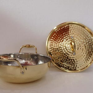 May include: A gold-coloured cooking pot with a matching lid. The pot has two handles and a hammered texture on the exterior. The lid is round with a similar hammered design. The pot and lid are on a white surface.