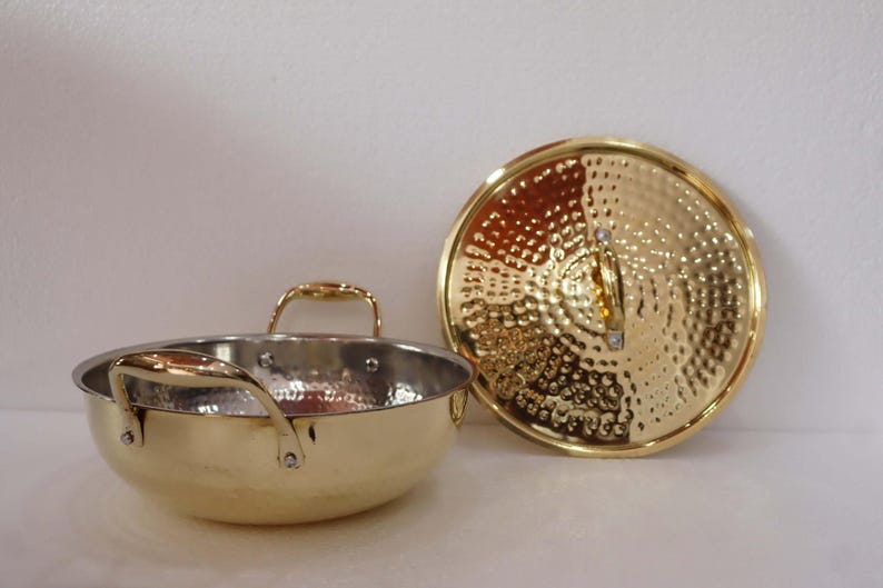May include: A gold-coloured cooking pot with a matching lid. The pot has two handles and a hammered texture on the exterior. The lid is round with a similar hammered design. The pot and lid are on a white surface.