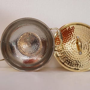 May include: A silver-coloured metal cooking pot with two handles and a gold-coloured lid. The pot and lid have a hammered texture. The lid is round with a handle in the centre. The pot is open, revealing the interior.