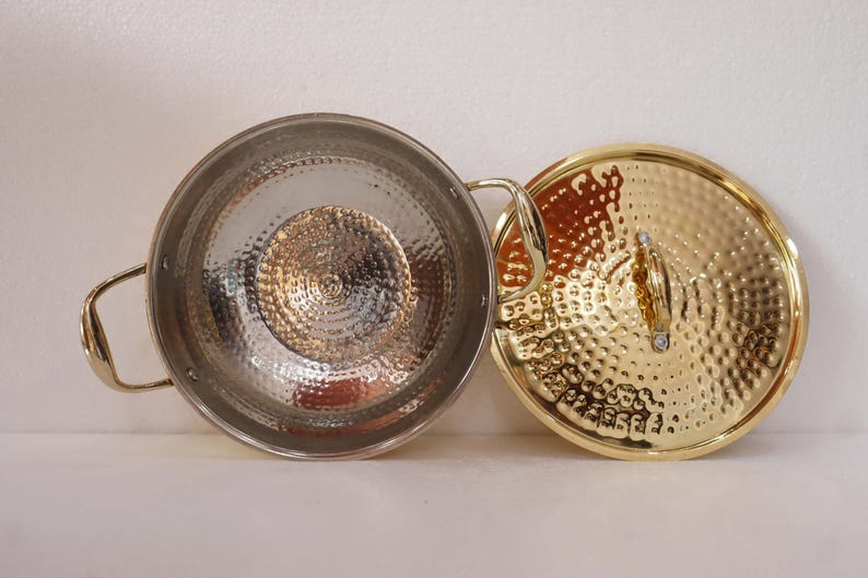 May include: A silver-coloured metal cooking pot with two handles and a gold-coloured lid. The pot and lid have a hammered texture. The lid is round with a handle in the centre. The pot is open, revealing the interior.