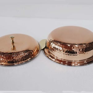 May include: Two copper serving dishes with lids. The dishes have a hammered texture and a shiny finish. The lid has a small handle. The dishes have brass handles. The dishes are likely for serving food.