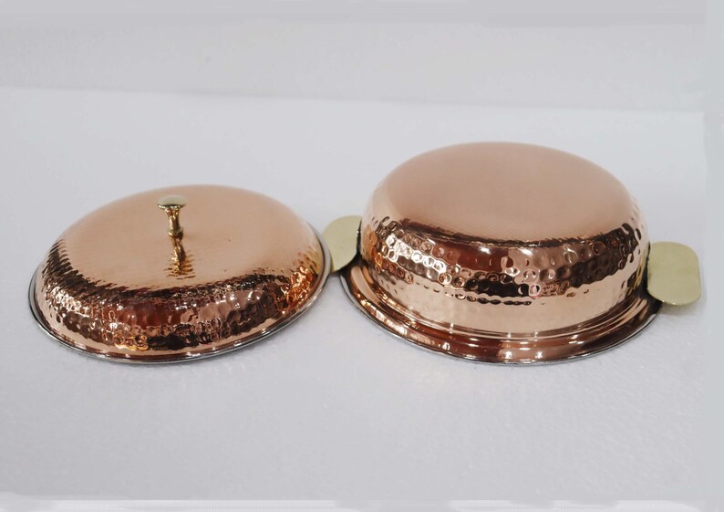 May include: Two copper serving dishes with lids. The dishes have a hammered texture and a shiny finish. The lid has a small handle. The dishes have brass handles. The dishes are likely for serving food.