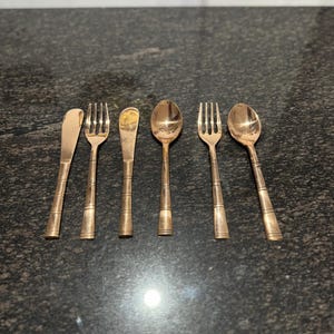 May include: A set of six antique gold-colored flatware pieces, including forks, spoons, and butter knives. The utensils are arranged on a dark, speckled surface, showcasing their vintage design and polished finish. Ideal for a vintage-themed dining setting.