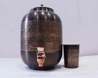 Könnte beinhalten: Ein kupferfarbener Wasserspender mit einem verzierten floralen Muster und einem Wasserhahn. Ein passender Becher befindet sich neben dem Spender. Der Spender hat einen Deckel und einen kupferfarbenen Wasserhahn. Der Becher hat das gleiche florale Muster.