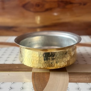 May include: A hammered brass bowl with a silver rim. The bowl has a wide, shallow shape and a textured surface. The brass has a warm, golden color, and the bowl appears to be handmade. The bowl is likely used for serving or display.