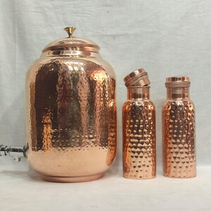 Op de afbeelding: Drie koperen waterflessen met gehammerde texturen. De grootste fles heeft een tuit en een deksel. De twee kleinere flessen hebben deksels.
