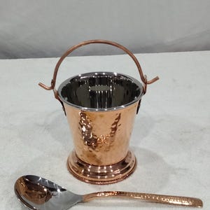 May include: A hammered copper serving bucket with a handle and a matching hammered copper spoon. The bucket has a stainless steel interior.