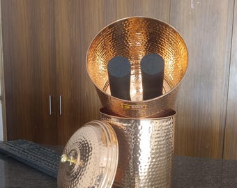 Può includere: Un filtro per l'acqua in rame con una texture martellata. Il filtro comprende una base cilindrica, una sezione superiore a forma di ciotola e un coperchio. All'interno della ciotola sono visibili due filtri cilindrici neri. Il filtro ha un rubinetto e un pomello dorato sul coperchio.