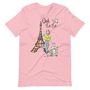 Op de afbeelding: Roze T-shirt met een afbeelding van de Eiffeltoren, een vrouw met een koffer en de tekst "Ooh La La" met een papieren vliegtuig.