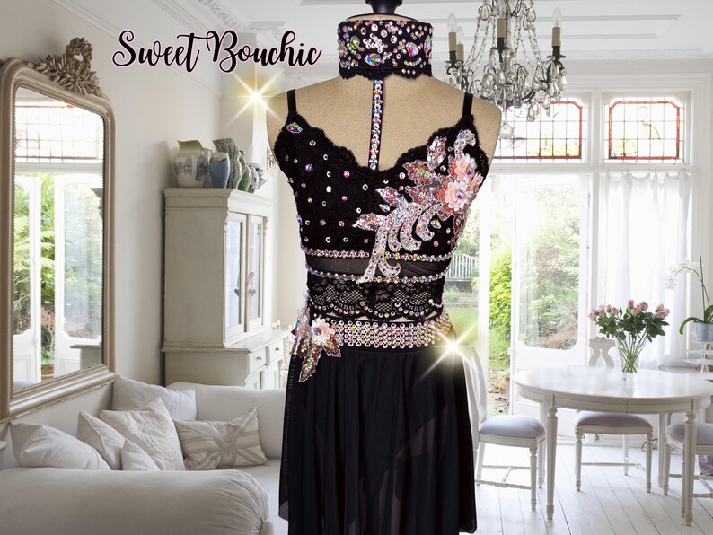 May include: A black dance costume with a sheer top and skirt. The top features a floral design with rhinestones and a black choker with rhinestones. The skirt is a simple black skirt with a sheer overlay.