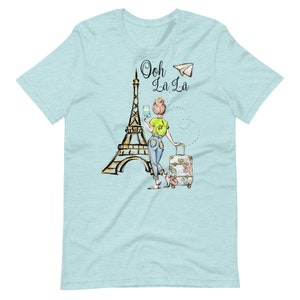 Op de afbeelding: Een lichtblauw T-shirt met een afbeelding van een vrouw die voor de Eiffeltoren staat met een koffer en een papieren vliegtuig. De tekst "Ooh La La" staat op het shirt gedrukt.