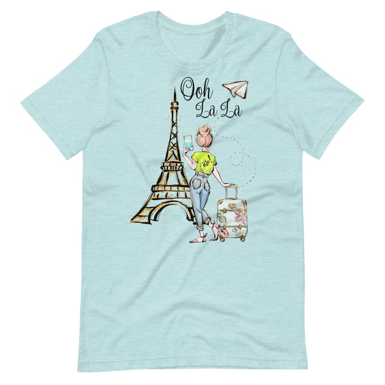 Op de afbeelding: Een lichtblauw T-shirt met een afbeelding van een vrouw die voor de Eiffeltoren staat met een koffer en een papieren vliegtuig. De tekst "Ooh La La" staat op het shirt gedrukt.