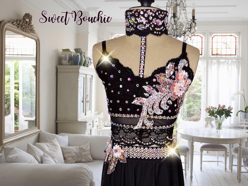 May include: Black dance costume with a black lace bodice, a black skirt, and a rhinestone belt. The bodice has a floral design with pink and peach accents. The costume is decorated with rhinestones.