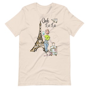 Op de afbeelding: Een beige t-shirt met een afbeelding van een vrouw die voor de Eiffeltoren staat met een koffer en een smartphone. De tekst "Ooh La La" staat boven de Eiffeltoren.